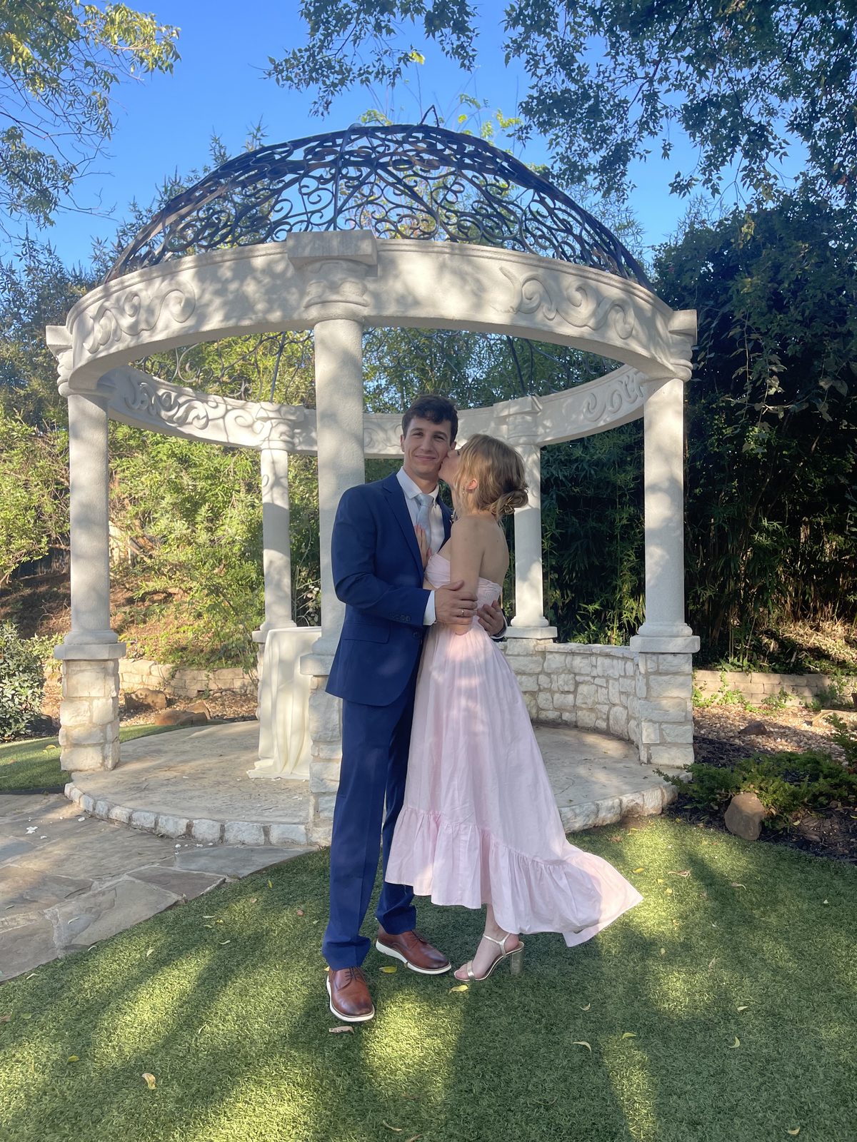 Francesca in a pink dress kissing Simon by a white gazebo at the third wedding they attended together